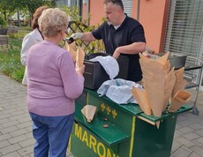 An einem Maronistand werden frisch gebratene Maroni in Papiertüten ausgegeben, während mehrere Personen im Freien auf der Terrasse zusammenkommen.