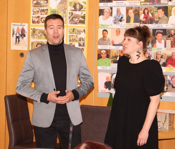 Eine Dame und ein Herr aus dem Künstlerkollegium Manfred Grössler stehen nebeneinander und singen gemeinsam vor den Bewohnerinnen und Bewohnern des Caritas Pflegewohnhauses Graz-Straßgang. Beide wirken konzentriert und herzlich, während sie dem Publikum mit ihrer Darbietung eine Freude bereiten. Im Vordergrund sind einige Zuhörer*innen zu sehen, die aufmerksam lauschen.
