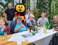 Bewohner*innen in Rollstühlen sitzen gemeinsam mit Mitarbeiter*innen an einem Tisch. Mit dabei ist ein Bienen-Maskottchen, das für fröhliche Stimmung sorgt.