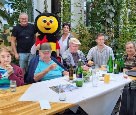 Bewohner*innen in Rollstühlen sitzen gemeinsam mit Mitarbeiter*innen an einem Tisch. Mit dabei ist ein Bienen-Maskottchen, das für fröhliche Stimmung sorgt.