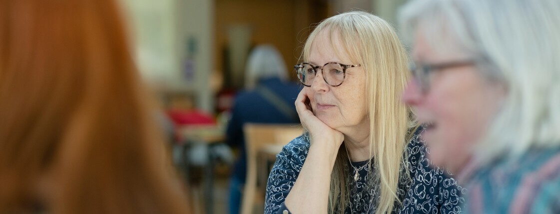 Drei Teilnehmerinnen einer Selbsthilfegruppe sitzen mit Kaffee an einem Tisch und hören einer anderen Seniorin zu.