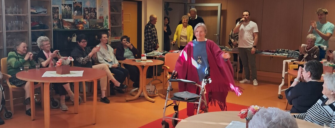 Eine Person präsentiert mit einer Gehhilfe auf einem roten Laufsteg eine Modeschau im barrierefreien Gemeinschaftsraum des Pflegewohnhauses, während das Publikum ringsum an Tischen sitzt.