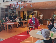 Eine Person präsentiert mit einer Gehhilfe auf einem roten Laufsteg eine Modeschau im barrierefreien Gemeinschaftsraum des Pflegewohnhauses, während das Publikum ringsum an Tischen sitzt.