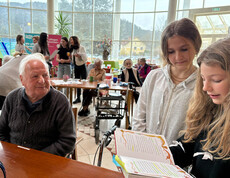 Schüler*innen lesen Senior*innen im Gemeinschaftsraum von Haus Suavitas aus Büchern vor, mehrere Tische sind besetzt.