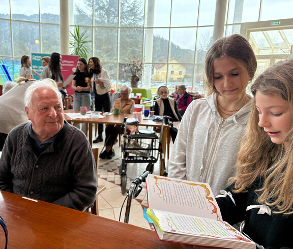 Schüler*innen lesen Senior*innen im Gemeinschaftsraum von Haus Suavitas aus Büchern vor, mehrere Tische sind besetzt.
