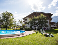 In einem großen Garten hinter einem Gasthaus ist eine große Poolanlage. Zwei Liegestühle stehen auf der Wiese.
