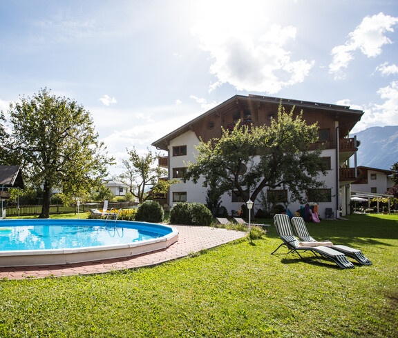 In einem großen Garten hinter einem Gasthaus ist eine große Poolanlage. Zwei Liegestühle stehen auf der Wiese.