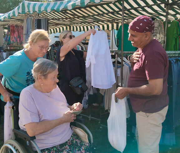 Seniorin im Rollstuhl zahlt an einem Kleidungsstand, begleitet von zwei Frauen