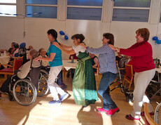 Pflegekräfte und Besucher*innen tanzen in Polonaise durch den Saal, auch Rollstuhlnutzende sind dabei