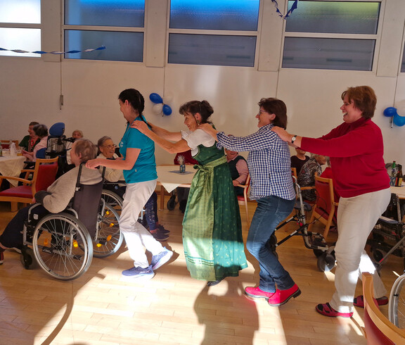 Pflegekräfte und Besucher*innen tanzen in Polonaise durch den Saal, auch Rollstuhlnutzende sind dabei
