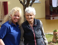 Eine Bewohnerin und eine Mitarbeiterin lächeln in die Kamera. Im Hintergrund ist das Pflegewohnhaus zu sehen.