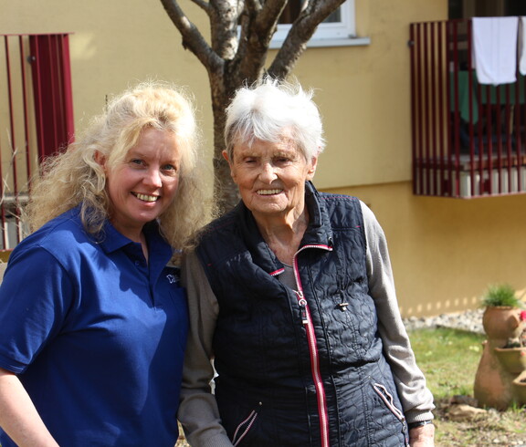 Eine Bewohnerin und eine Mitarbeiterin lächeln in die Kamera. Im Hintergrund ist das Pflegewohnhaus zu sehen.