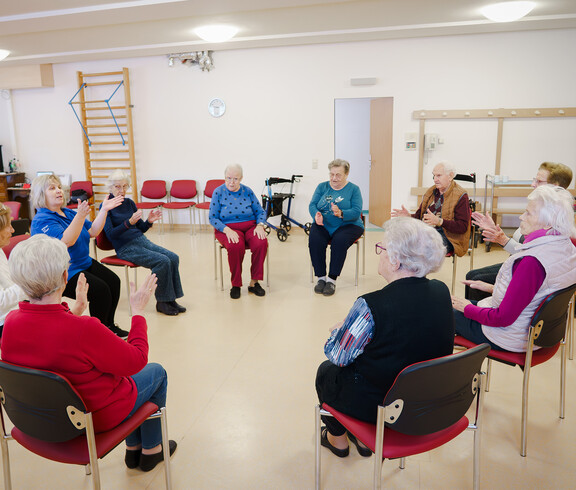 Senior*innen sitzen im Kreis und machen angeleitete Bewegungsübungen.