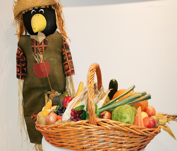 Eine herbstliche Dekoration mit einer Vogelscheuche, die neben einem kleinen Tisch steht. Auf dem Tisch befindet sich ein Korb voller frischer Ernteprodukte wie Kürbisse, Maiskolben und Äpfel. Rund um den Tisch liegen weitere Kürbisse, was eine festliche Stimmung passend zum Erntedankfest schafft.