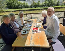 Mehrere Bewohnerinnen und Bewohner sitzen gemeinsam auf einer Bierbank, genießen Kaffee und Kuchen und verbringen gesellige Stunden beim Ausflug unseres Pflegewohnhauses.