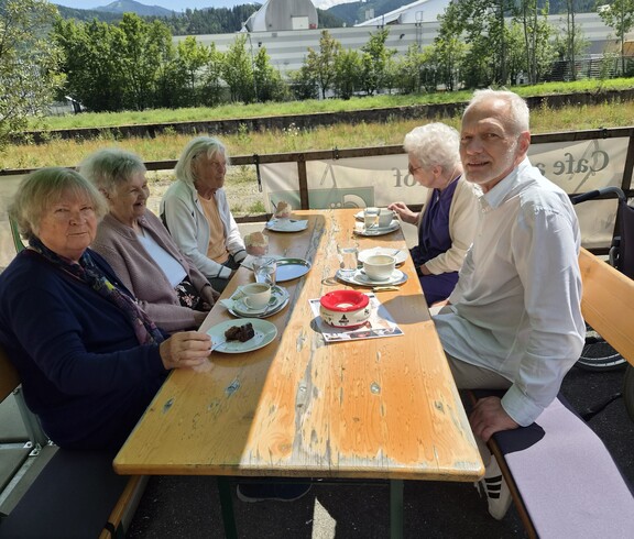 Mehrere Bewohnerinnen und Bewohner sitzen gemeinsam auf einer Bierbank, genießen Kaffee und Kuchen und verbringen gesellige Stunden beim Ausflug unseres Pflegewohnhauses.