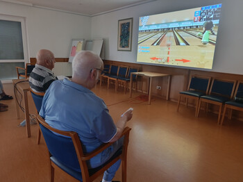 Bewohner*innen spielen Wii Bowling im Gemeinschaftsraum und verfolgen das Spiel auf einer großen Projektion.