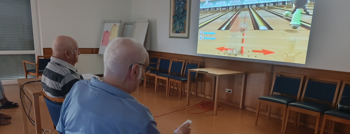 Bewohner*innen spielen Wii Bowling im Gemeinschaftsraum und verfolgen das Spiel auf einer großen Projektion.