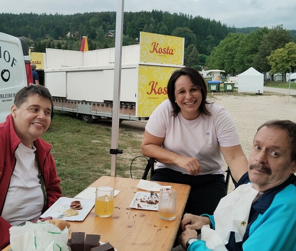 Zwei Männer und eine Frau sitzen an Holztisch mit Kuchen und Getränken im Freien