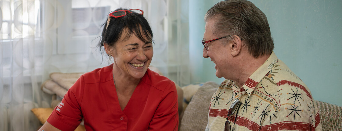 Eine Heimhelferin sitzt mit einem älteren Herrn am Esstisch und schreibt mit ihm die Einkaufsliste.