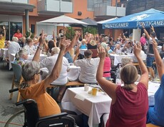 Beim Sommerfest im Innenhof des Pflegewohnhauses sitzen zahlreiche Bewohnerinnen und Bewohner an festlich gedeckten Tischen und heben gemeinsam die Arme, während das Musikprogramm für Unterhaltung sorgt.