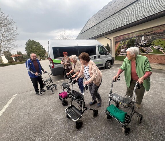 Eine Gruppe von Bewohnerinnen des Pflegewohnhauses ist gemeinsam mit Begleitpersonen auf dem Weg zum Café. Die Gruppe steht vor einem Transporter und bereitet sich mit Rollatoren auf den kurzen Fußweg vor.