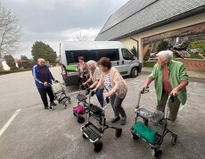 Eine Gruppe von Bewohnerinnen des Pflegewohnhauses ist gemeinsam mit Begleitpersonen auf dem Weg zum Café. Die Gruppe steht vor einem Transporter und bereitet sich mit Rollatoren auf den kurzen Fußweg vor.
