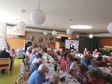 Beim Sommerfest im Pflegewohnhaus haben sich zahlreiche Bewohnerinnen und Bewohner, Angehörige sowie Mitarbeitende in einem festlich geschmückten Gemeinschaftsraum versammelt, genießen Speisen und Getränke an langen Tischen und verfolgen gemeinsam das Unterhaltungsprogramm.