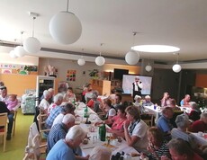 Beim Sommerfest im Pflegewohnhaus haben sich zahlreiche Bewohnerinnen und Bewohner, Angehörige sowie Mitarbeitende in einem festlich geschmückten Gemeinschaftsraum versammelt, genießen Speisen und Getränke an langen Tischen und verfolgen gemeinsam das Unterhaltungsprogramm.
