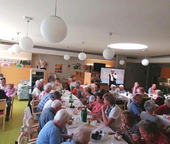Beim Sommerfest im Pflegewohnhaus haben sich zahlreiche Bewohnerinnen und Bewohner, Angehörige sowie Mitarbeitende in einem festlich geschmückten Gemeinschaftsraum versammelt, genießen Speisen und Getränke an langen Tischen und verfolgen gemeinsam das Unterhaltungsprogramm.