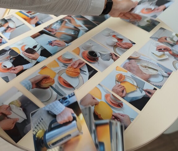 Auf einem hellen Tisch liegen zahlreiche Fotokarten verteilt. Sie zeigen Hände bei alltäglichen Handgriffen wie Kaffee einschenken, Zucker umrühren, einen Kuchen halten oder Geschirr anfassen. Von oben greift eine Hand mit Armbanduhr nach einer der Karten, um sie zu ordnen.