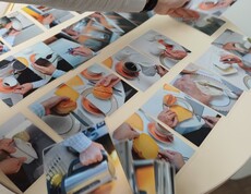 Auf einem hellen Tisch liegen zahlreiche Fotokarten verteilt. Sie zeigen Hände bei alltäglichen Handgriffen wie Kaffee einschenken, Zucker umrühren, einen Kuchen halten oder Geschirr anfassen. Von oben greift eine Hand mit Armbanduhr nach einer der Karten, um sie zu ordnen.
