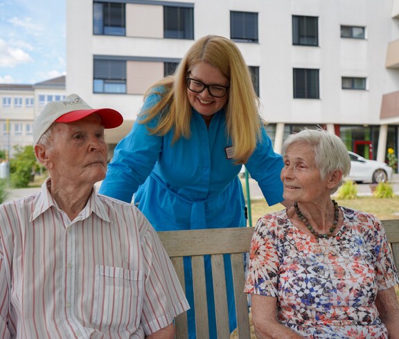 Zwei Bewohner*innen und die Pflegeleitung sitzen draußen auf einer Bank.