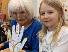 Im Pflegewohnhaus steht ein Kind neben einer Bewohnerin im Rollstuhl und präsentiert gemeinsam ein liebevoll gestaltetes Bastelwerk im hellen Gemeinschaftsraum.