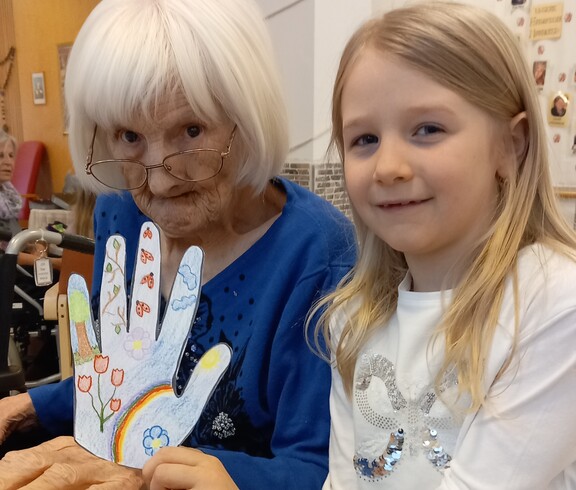 Im Pflegewohnhaus steht ein Kind neben einer Bewohnerin im Rollstuhl und präsentiert gemeinsam ein liebevoll gestaltetes Bastelwerk im hellen Gemeinschaftsraum.