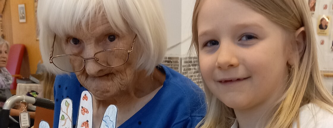 Im Pflegewohnhaus steht ein Kind neben einer Bewohnerin im Rollstuhl und präsentiert gemeinsam ein liebevoll gestaltetes Bastelwerk im hellen Gemeinschaftsraum.