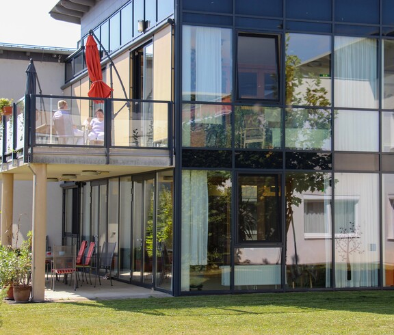 Außenansicht des Hauses Lisa mit großen Glasfronten und einem Balkon, auf dem zwei Personen sitzen. Zu sehen ist auch eine Grünfläche.