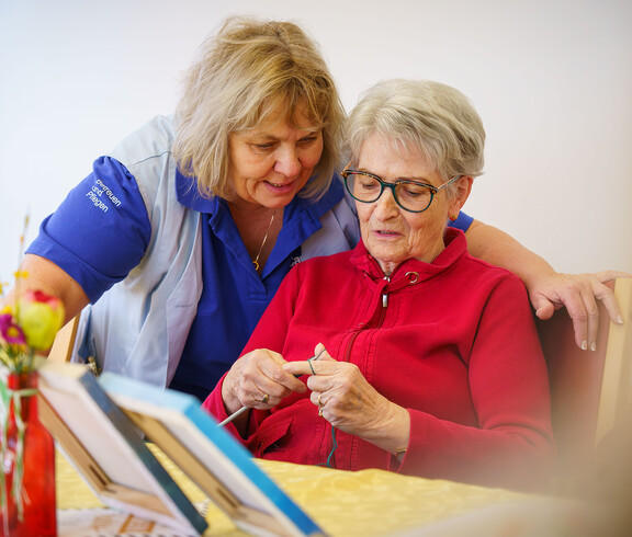 Betreuungskraft unterstützt Seniorin beim Stricken am Tisch
