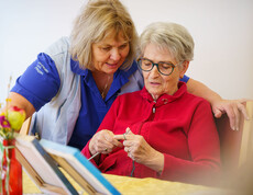 Betreuungskraft unterstützt Seniorin beim Stricken am Tisch