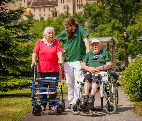 Ein Pfleger steht mit einer Dame am Rollator und einem Mann im Rollstuhl im Garten des Hauses und lächelt die Dame an.