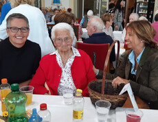 Eine Seniorin in roter Jacke sitzt mit zwei Frauen am Tisch, umgeben von Getränken und herbstlicher Deko.