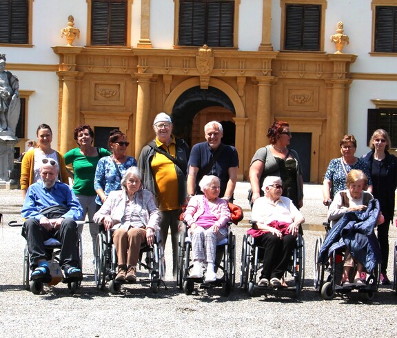 Einige Bewohner*innen sitzen in Rollstühlen nebeneinander. Hinter ihnen befinden sich Pflegepersonal sowie Angehörige. Im Hintergrund ist das Schloss Eggenberg zu sehen.