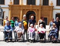 Einige Bewohner*innen sitzen in Rollstühlen nebeneinander. Hinter ihnen befinden sich Pflegepersonal sowie Angehörige. Im Hintergrund ist das Schloss Eggenberg zu sehen.