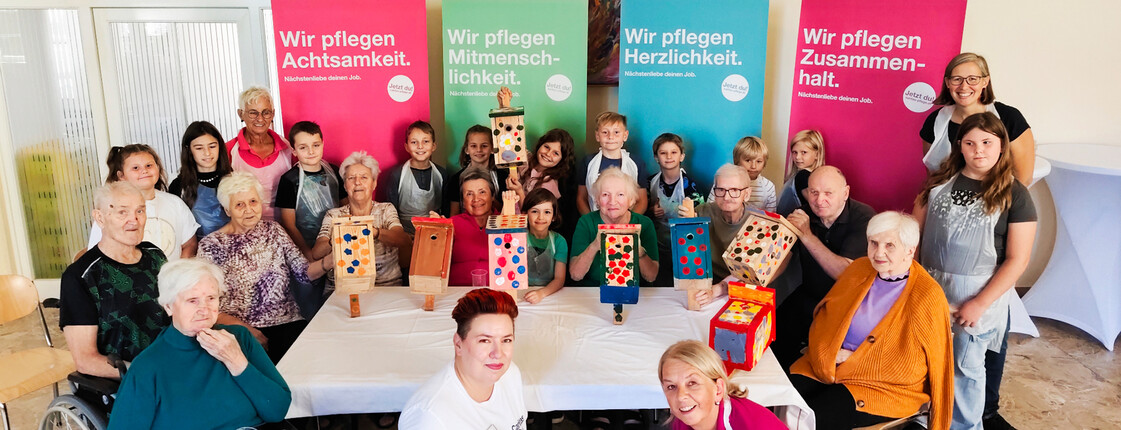 Jung und Alt gestalten Vogelbrutkästen gemeinsam Senior*innen und Kinder posieren vor bunten Rollups mit selbstgebauten Vogelbrutkästen.