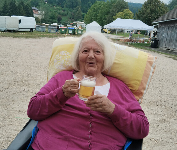 Ältere Frau im Rollstuhl hält Bierglas und blickt vergnügt in die Kamera