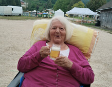 Ältere Frau im Rollstuhl hält Bierglas und blickt vergnügt in die Kamera