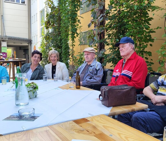 Mehrere Bewohner*innen in Rollstühlen sitzen gemeinsam mit Angehörigen und Pflegepersonal an einem Tisch. Sie befinden sich im Freien am Marktplatz Wies und scheinen einer Darbietung zuzusehen, die außerhalb des Bildes stattfindet.