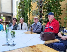 Mehrere Bewohner*innen in Rollstühlen sitzen gemeinsam mit Angehörigen und Pflegepersonal an einem Tisch. Sie befinden sich im Freien am Marktplatz Wies und scheinen einer Darbietung zuzusehen, die außerhalb des Bildes stattfindet.