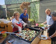 Beim Sommerfest im Pflegewohnhaus wird im Innenhof frisch gegrillt: Mehrere Personen bereiten an einer großen Grillplatte buntes Gemüse und Würstchen zu, während Gäste ihre Teller füllen und das gemeinsame Festessen genießen.