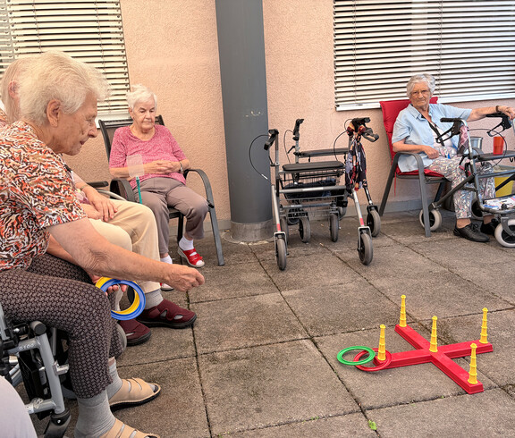 Bewohner*innen sitzen im Kreis und nehmen an einem Ringwurfspiel auf der Terrasse teil.